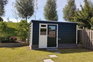 A small, dark-colored shed with a white door and window, surrounded by a grassy yard and a wooden fence. - Holiday home with 3 bedrooms (Wolphaartsdijk)