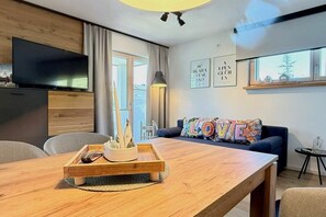 Ein modernes Wohnzimmer mit einem Holzesstisch, einer Couch mit bunten Kissen, einem an der Wand montierten Fernseher und einem Fenster mit Blick.