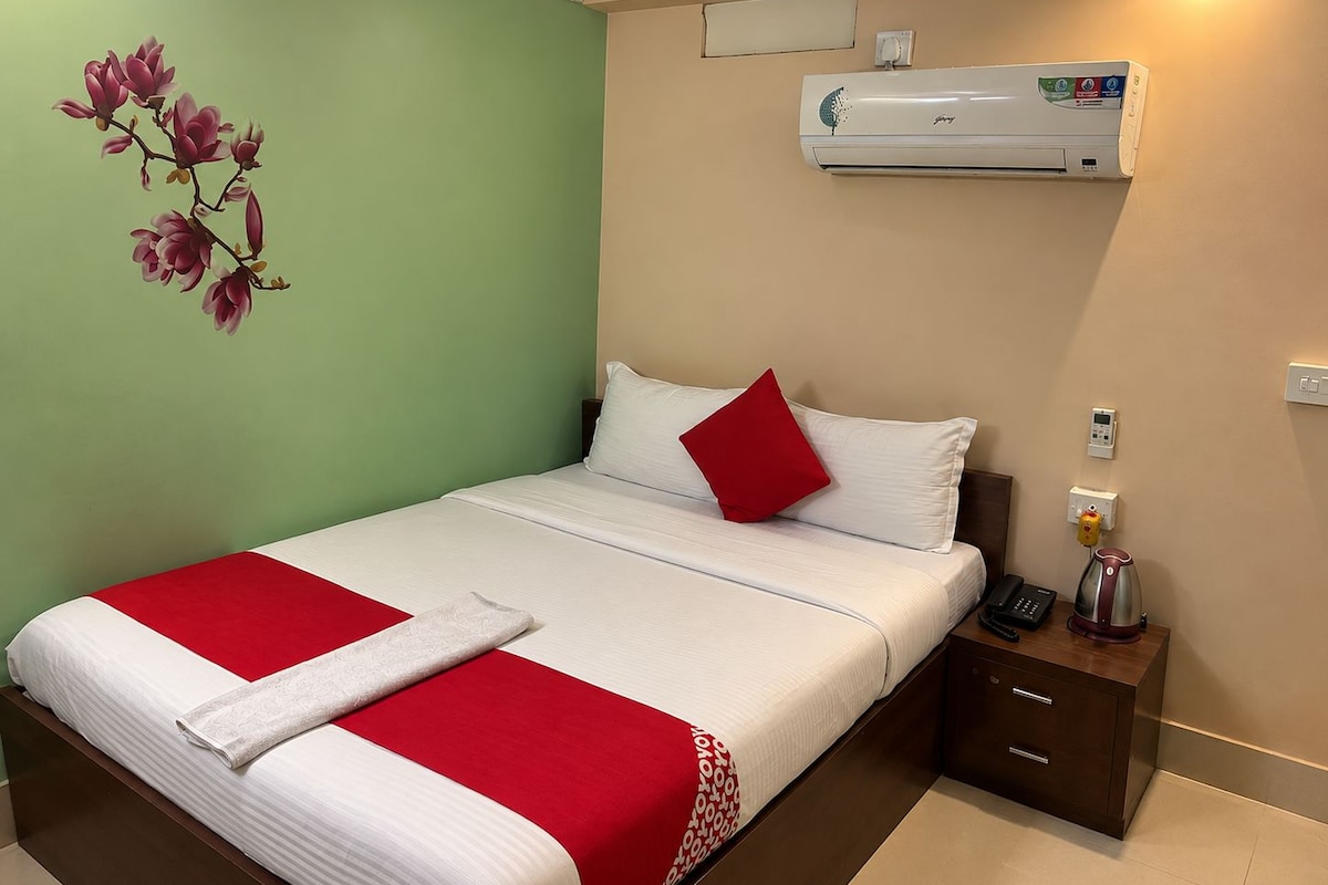 A hotel room with a bed, a red and white bedspread, a nightstand with a phone and a kettle, and an air conditioning unit on the wall.