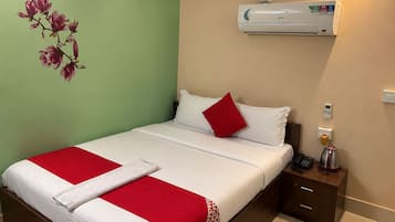 Habitación de hotel con una cama, colcha roja y blanca, mesita de noche con teléfono y hervidor, y unidad de aire acondicionado en la pared.