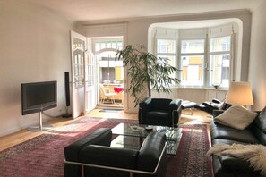 A living room with a flat-screen TV, black leather sofa, a glass coffee table, and a large potted plant. - Holiday apartment with 1 bedroom (Lindau)