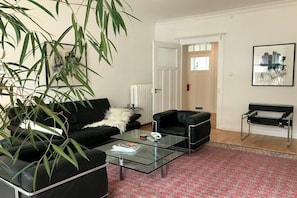 A living room with a black sofa, a glass coffee table, a red patterned rug, a black bench, and a large plant. - Holiday apartment with 1 bedroom (Lindau)