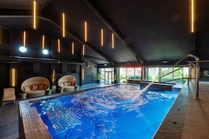 Une piscine intérieure à l’eau d’un bleu cristallin, entourée de chaises longues et dotée d’un plafond moderne au design épuré, agrémenté de luminaires linéaires.