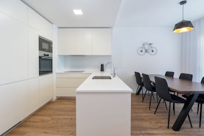 A modern kitchen with white cabinetry, a built-in oven, and a dining area with a wooden table and chairs. - Deluxe 1-Bedroom Apartment with Panoramic Pool - Happy Place in Faro (Faro)