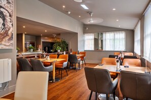 A modern dining area with wooden flooring, multiple tables and chairs, and a large window with sheer curtains. - Park Hall, 2 Bedroom Apartment (Charnock Richard)
