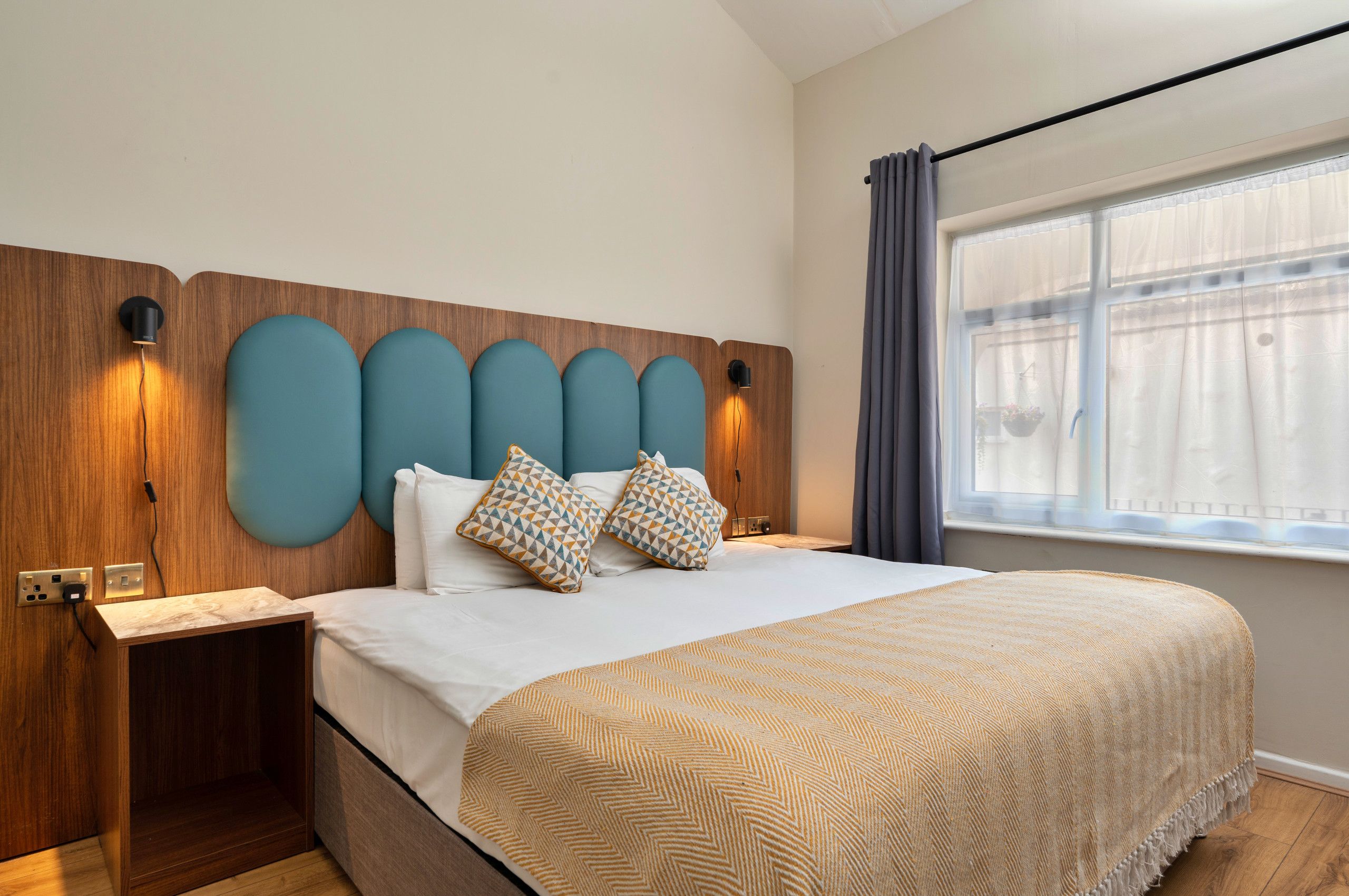 Une chambre d’hôtel comprenant un lit, une table de chevet, une tête de lit en bois avec des découpes circulaires, et une fenêtre avec des rideaux.