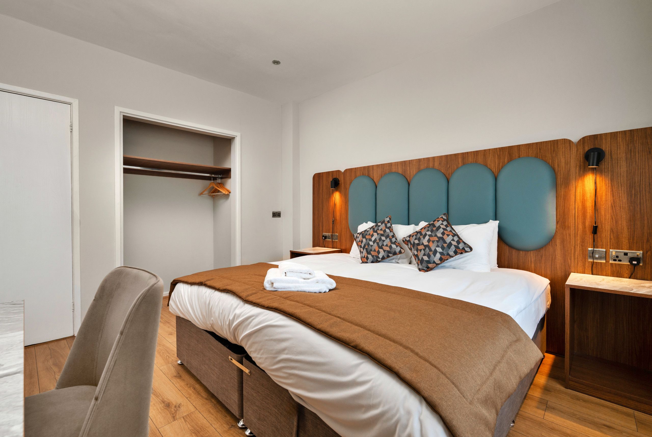 A hotel room with a bed, a chair, a wooden headboard, and a bedside table.