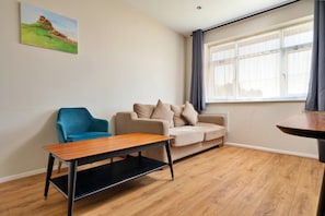 A modern living room with a blue armchair, a beige sofa with cushions, a wooden coffee table, and a window with curtains.