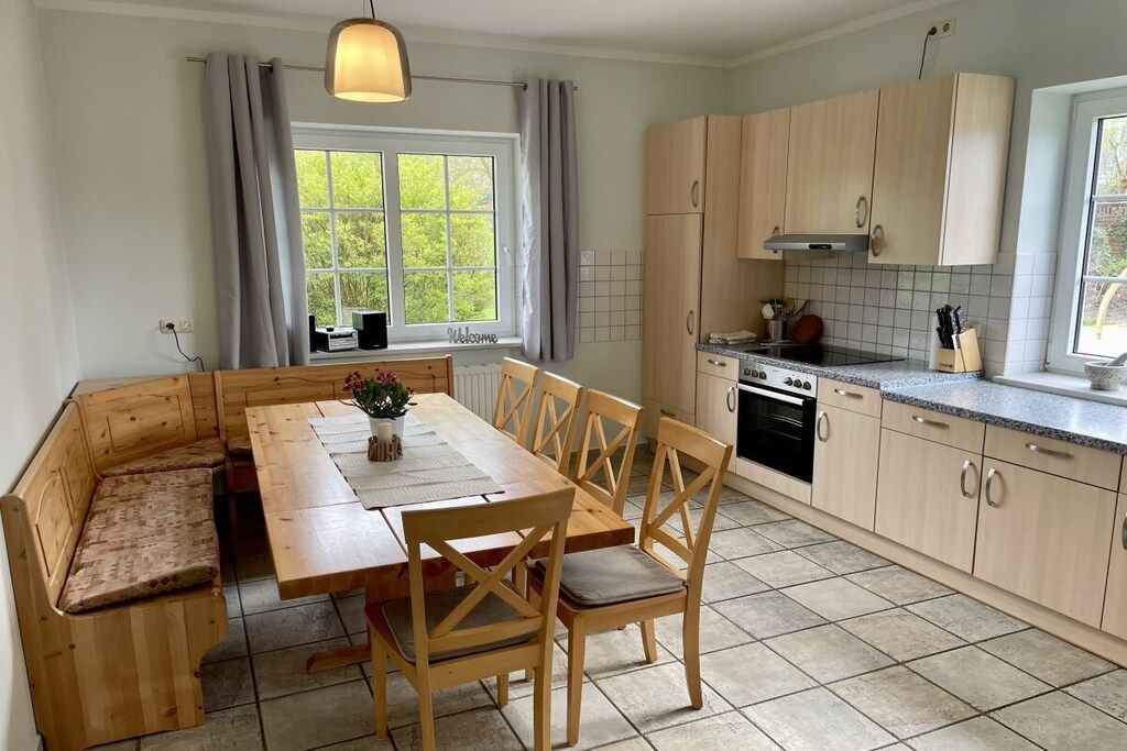 Una cocina con armarios de madera, una mesa de comedor con sillas, un horno empotrado y una ventana con cortinas.