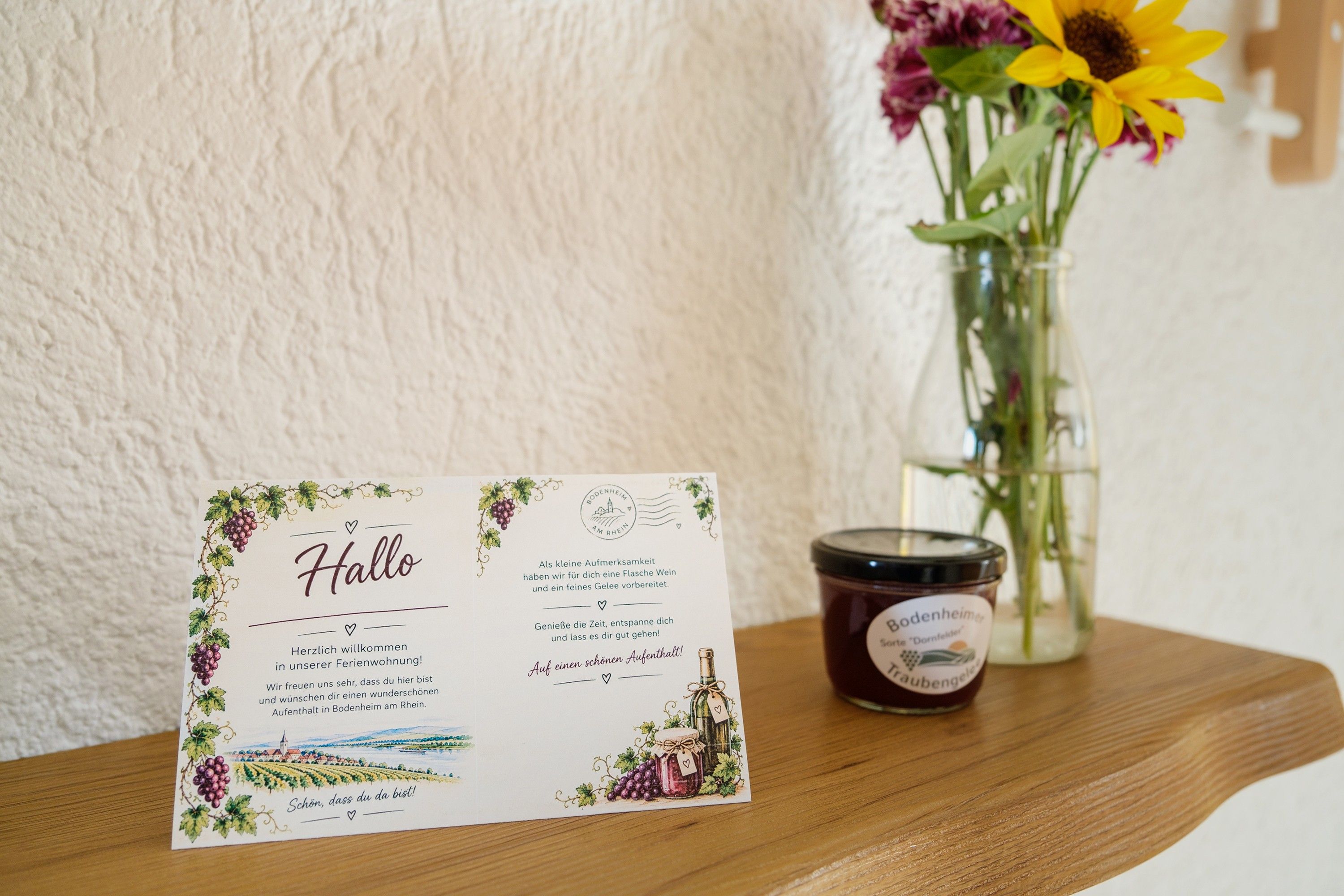 Una tarjeta de bienvenida con una ilustración de viñedo, una vela y un florero con flores sobre una mesa de madera.