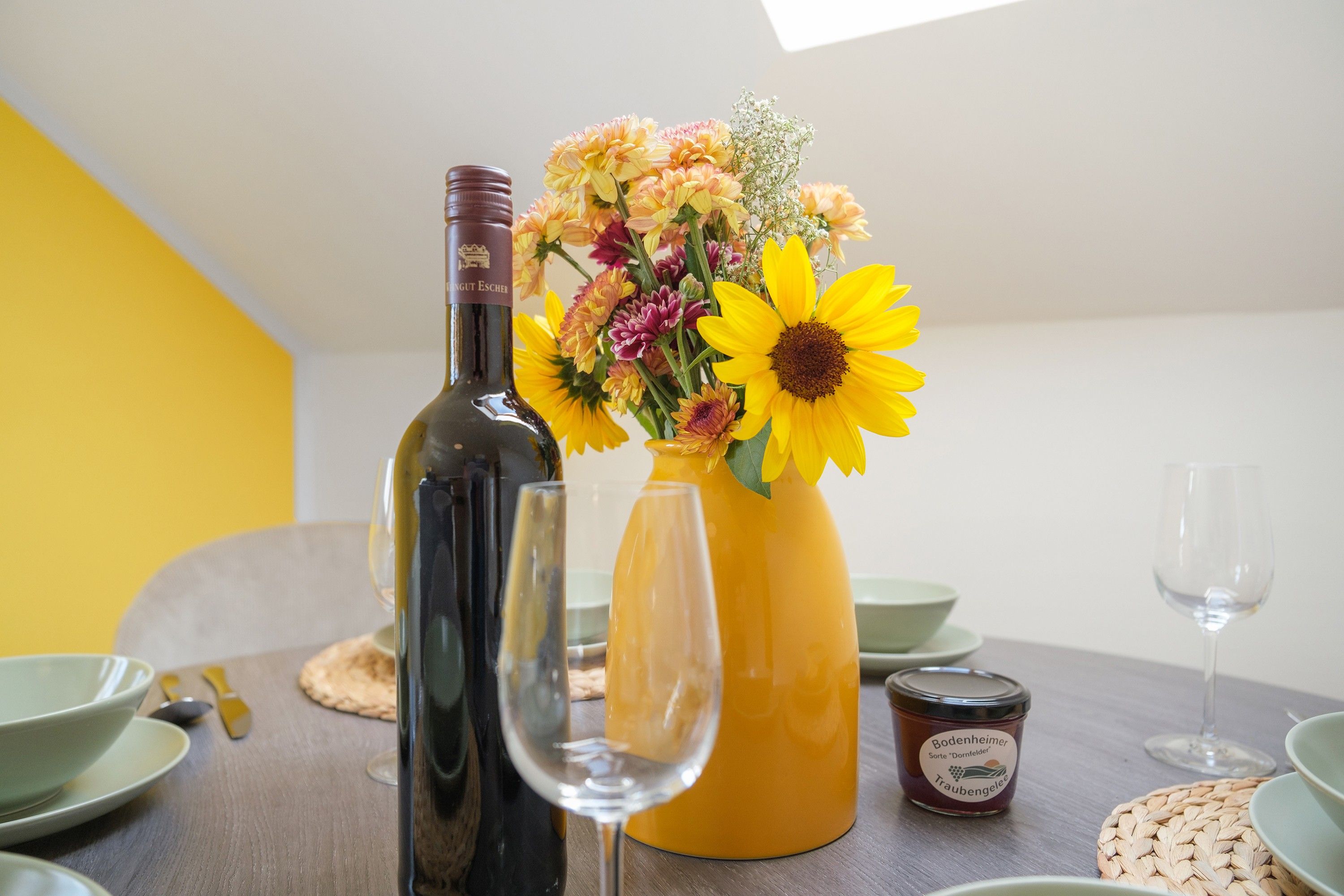 A dining table with a wine bottle, a glass, a vase with flowers, and a jar of jam.