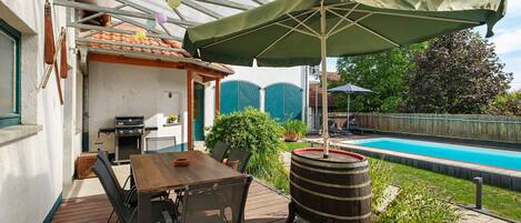 An outdoor patio with a wooden table and chairs, a barbecue grill, and a swimming pool in the background.