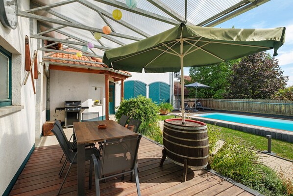 An outdoor patio with a wooden table and chairs, a barbecue grill, and a swimming pool in the background.