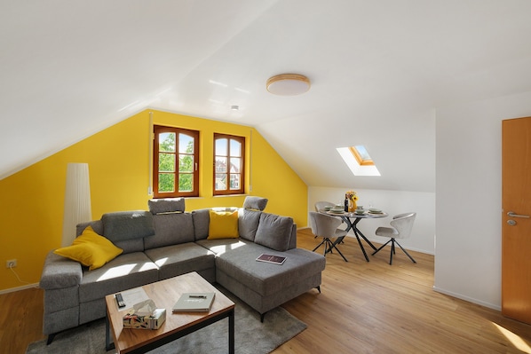 A modern living space with a sloped ceiling, a sectional sofa, a dining table with chairs, and a small kitchen area.