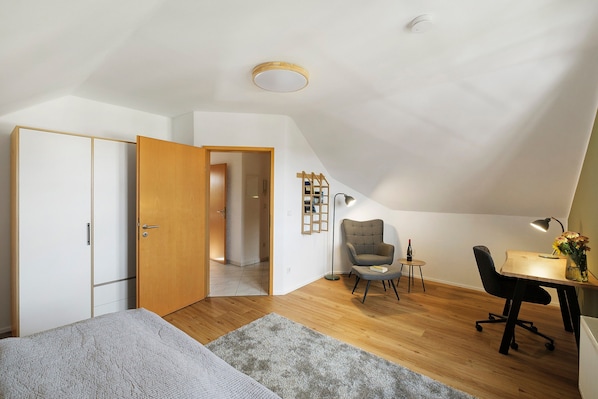 A modern bedroom with a sloped ceiling, a bed, a desk, a chair, and a small table with a lamp and a bottle.