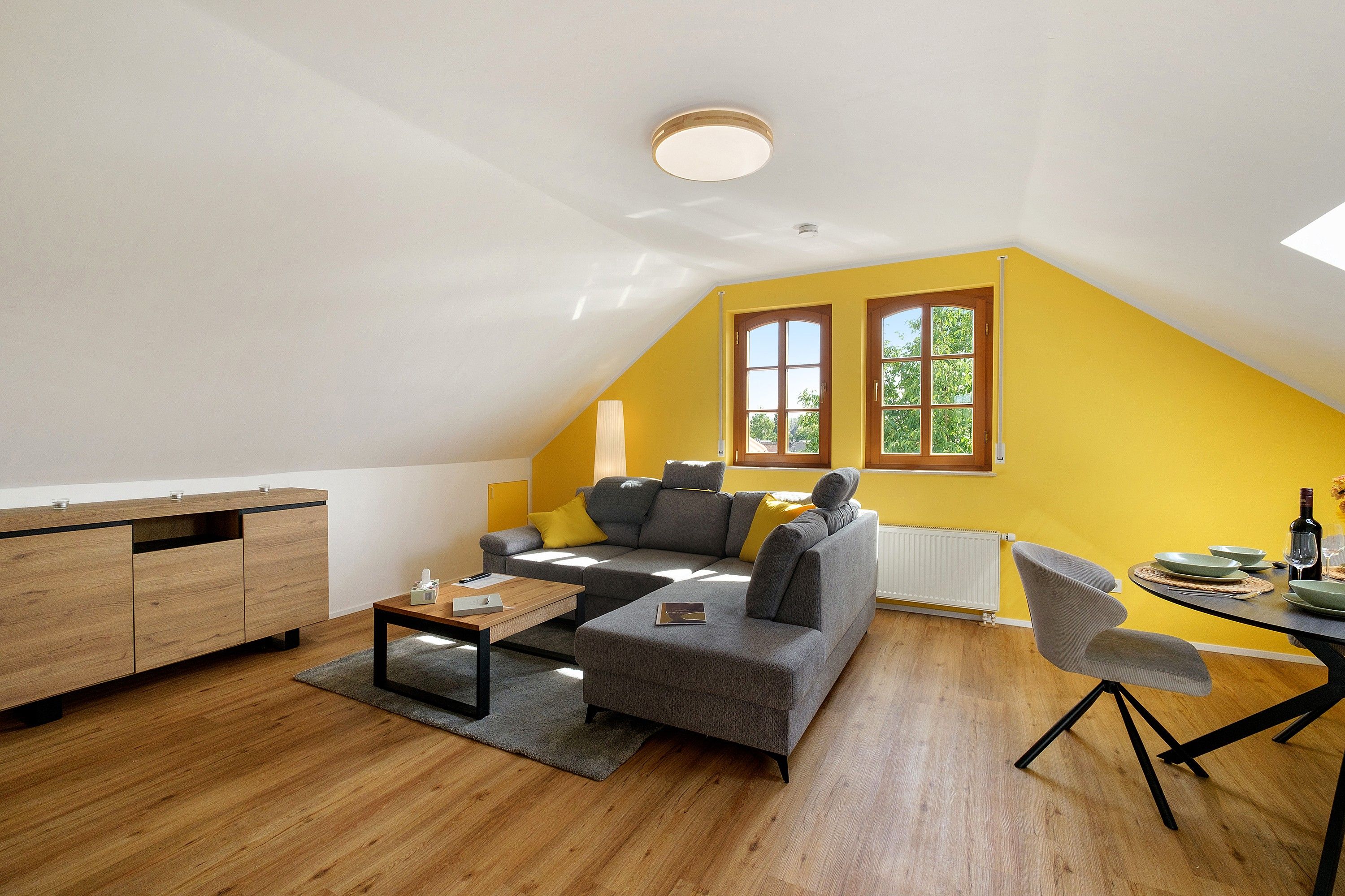Un moderno spazio abitativo in stile loft, con soffitto spiovente, un divano grigio, un tavolino in legno e un'area pranzo con tavolo imbandito per due persone.