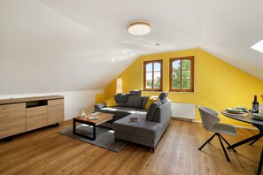 A modern loft-style living space with a sloped ceiling, a grey sofa, a wooden coffee table, and a dining area with a table set for two.