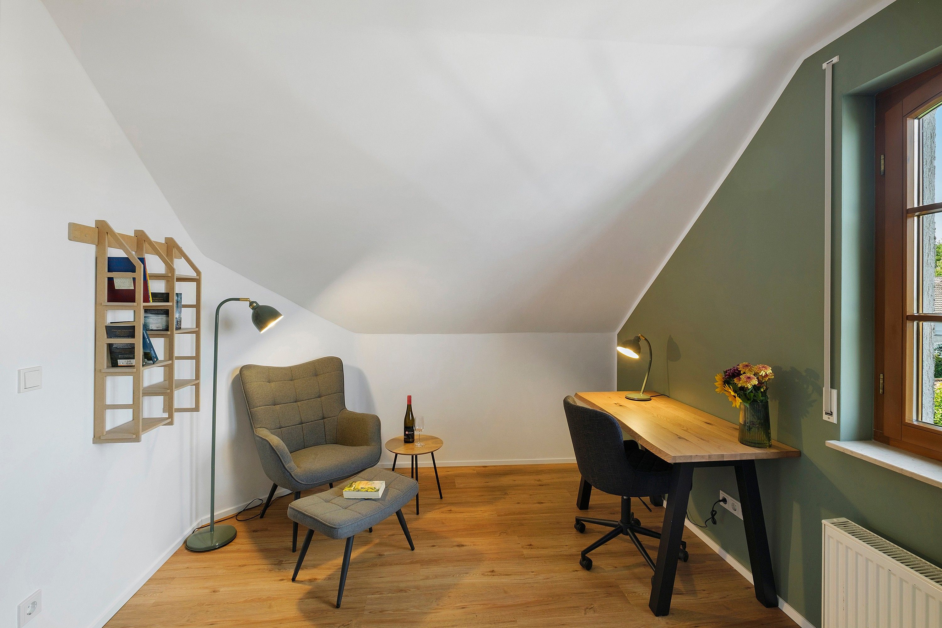 A loft-style room with a sloped ceiling, a wooden ladder bookshelf, a grey armchair, a small round table with a bottle and glass, a desk with a lamp, and a window with a view.