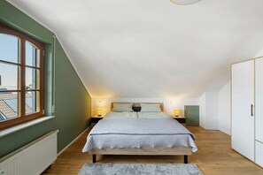 A bedroom with a sloped ceiling, a large window, a bed with a checkered bedspread, two bedside lamps, and a radiator.