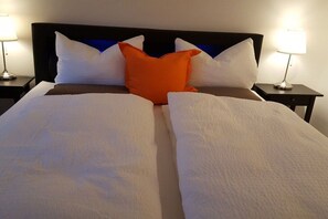 Ein Bett mit weißen Laken und einem einzelnen orangefarbenen Kissen, begleitet von zwei Nachttischen mit Lampen.