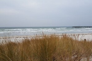 Ein Strand mit Wellen, einem Steg und hohem Gras im Vordergrund.