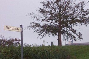 Ein Straßenschild mit der Aufschrift 'Werftstraße' in ländlicher Umgebung, mit einem großen Baum und einem Gebäude im Hintergrund.