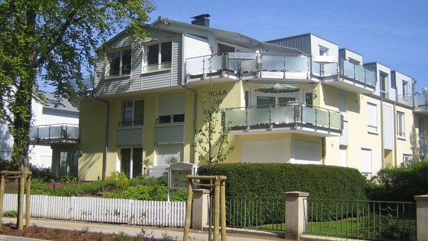 Un immeuble résidentiel à plusieurs étages, doté de balcons, d’une clôture en bois peinte en blanc et d’une haie bien entretenue.