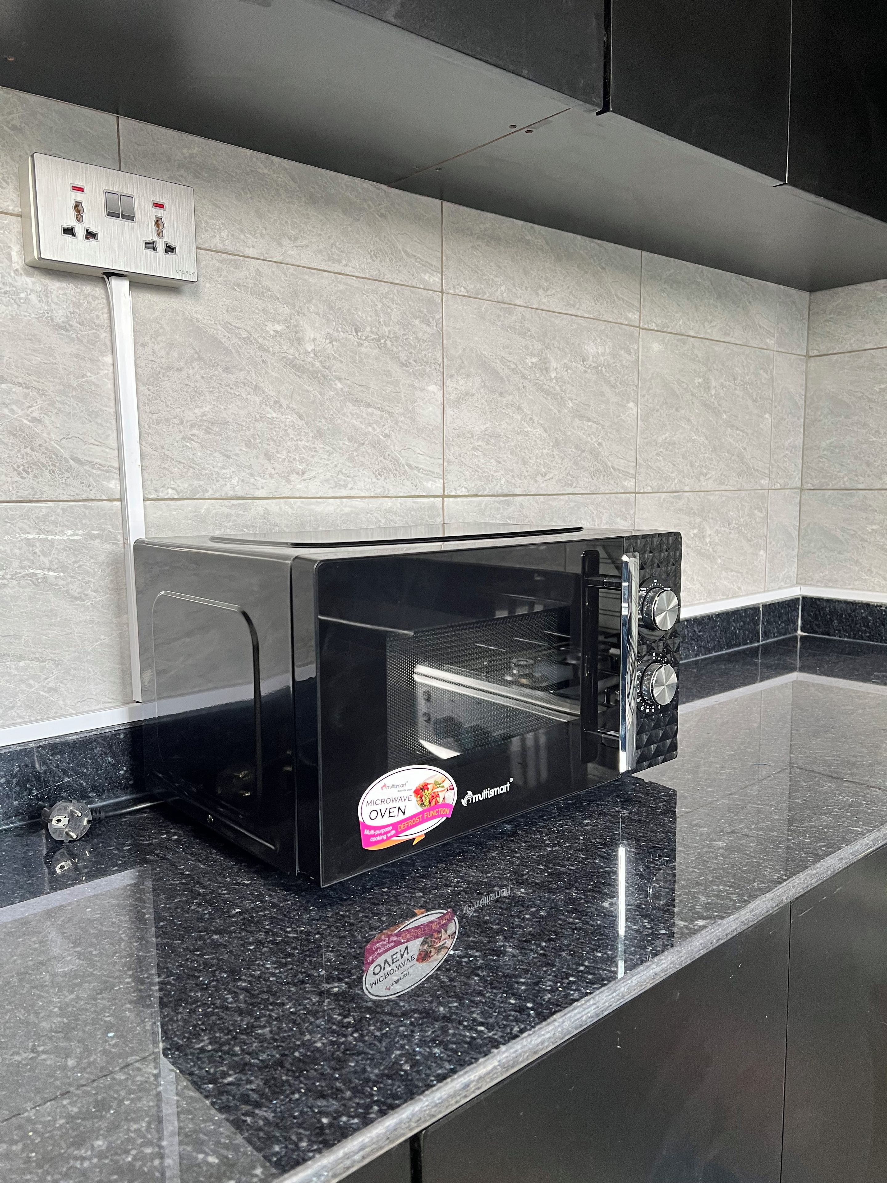 Eine schwarze Mikrowelle auf einer Arbeitsplatte aus Granit mit einer Edelstahl-Spritzschutzwand und einer an der Wand montierten Steckdose darüber.