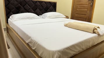 Un lit avec un cadre en bois et une tête de lit capitonnée, agrémenté de linge de lit blanc et d’une serviette roulée.