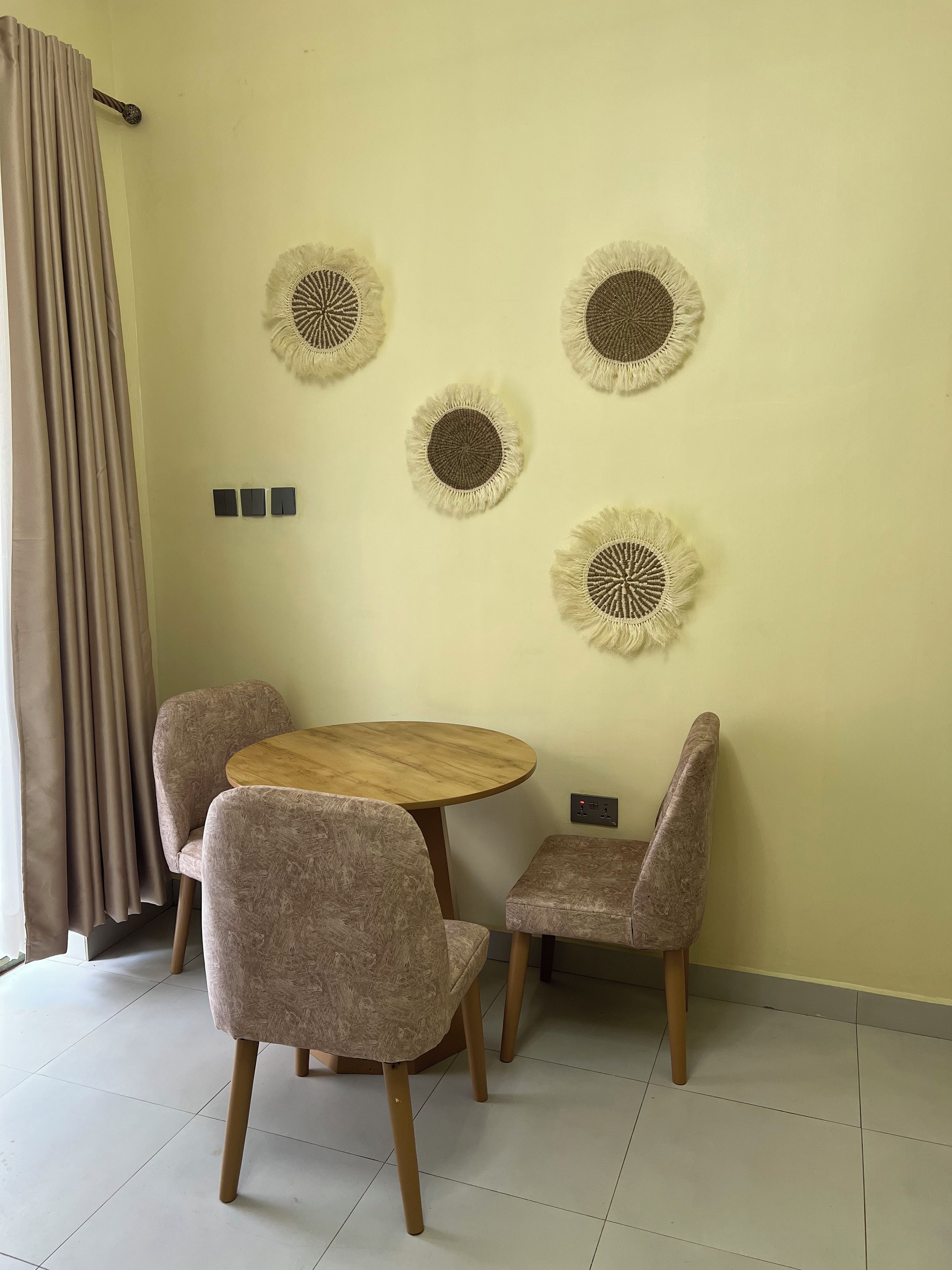 Une pièce avec une table ronde en bois et quatre chaises, une prise de courant fixée au mur et un rideau sur le côté gauche.
