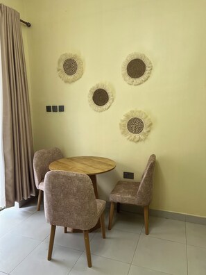 A room with a round wooden table and four chairs, a wall-mounted power outlet, and a curtain on the left.
