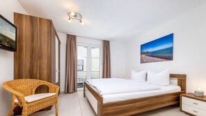 Ein Schlafzimmer mit Bett, Sessel, Nachttisch, Wand-Fernseher und einem Fenster mit Vorhängen.