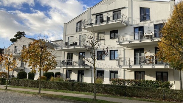 Ein mehrgeschossiges Wohngebäude mit Balkonen, einer gepflasterten Auffahrt und ausgewachsenen Bäumen.