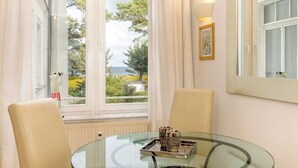 Ein Zimmer mit einem runden Glastisch, zwei Stühlen, einem Fenster mit Blick auf Bäume und ein Gebäude sowie einem gerahmten Bild an der Wand.