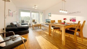 Un salon moderne avec un canapé noir, une table à manger en bois et des chaises. Un téléviseur est fixé au mur et une fenêtre laisse entrer la lumière naturelle.