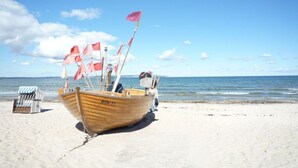 Ein hölzernes Boot mit der Kennung BIN-11 an einem sandigen Strand, ein gestreifter Liegestuhl und ein klarer Himmel.