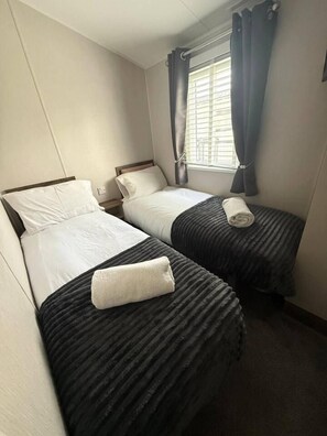 Habitación de hotel con dos camas individuales, una ventana con cortinas y una mesita de noche fijada a la pared.