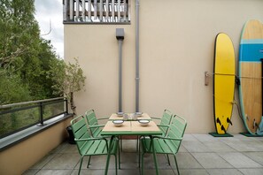 Une terrasse avec une table et des chaises, une rambarde et des planches de surf appuyées contre le mur.