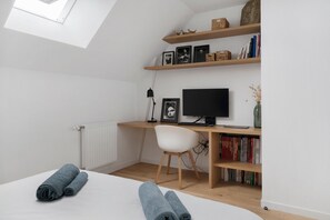 Une chambre avec un puits de lumière, un lit avec des serviettes roulées, un bureau avec un ordinateur, une chaise et des étagères à livres.