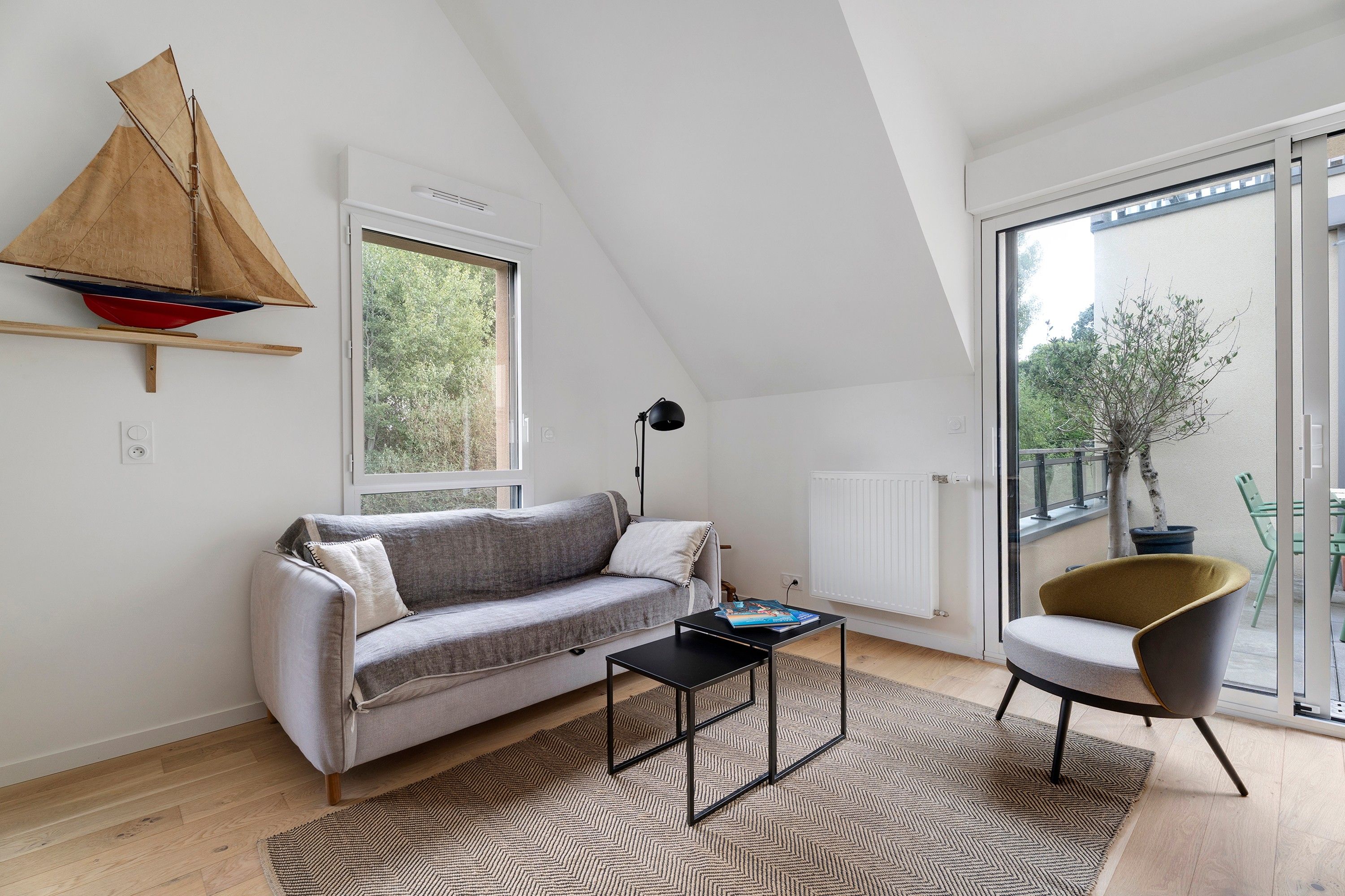 Une chambre de style loft moderne, avec un plafond en pente, une maquette de voilier, un canapé, une petite table, une chaise et une grande fenêtre donnant sur un paysage verdoyant.