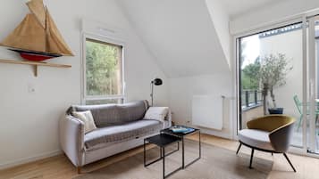 Une chambre de style loft moderne, avec un plafond en pente, une maquette de voilier, un canapé, une petite table, une chaise et une grande fenêtre donnant sur un paysage verdoyant.