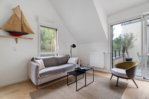 Une chambre de style loft moderne, avec un plafond en pente, une maquette de voilier, un canapé, une petite table, une chaise et une grande fenêtre donnant sur un paysage verdoyant.