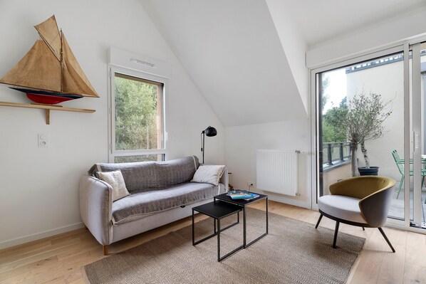 Een moderne loft slaapkamer met schuine wand, een model zeilboot, een bank, een klein tafeltje, een stoel en een groot raam met uitzicht op groen.