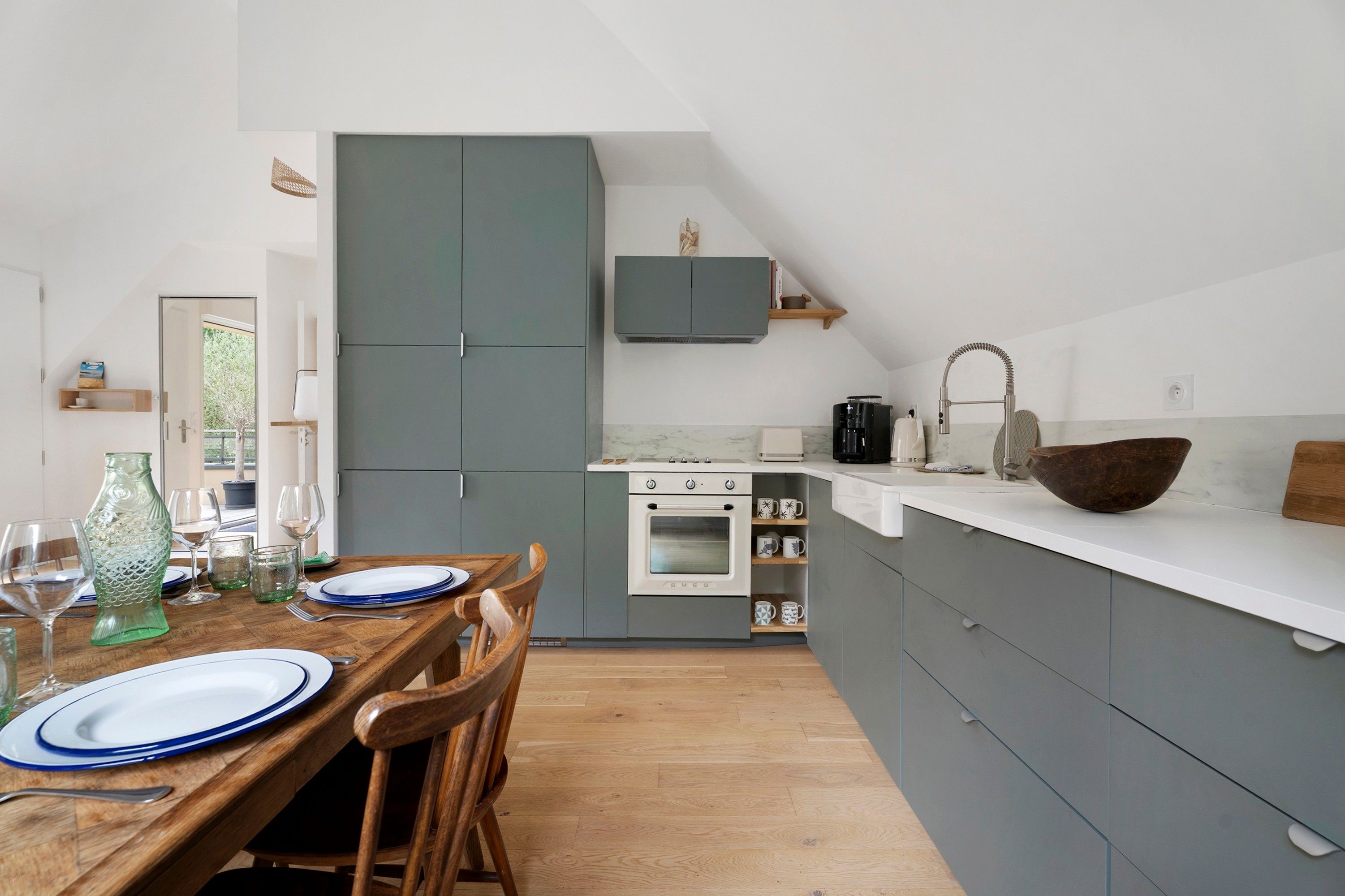 Une cuisine moderne avec des meubles gris, un plan de travail blanc et une table à manger en bois pour quatre personnes.