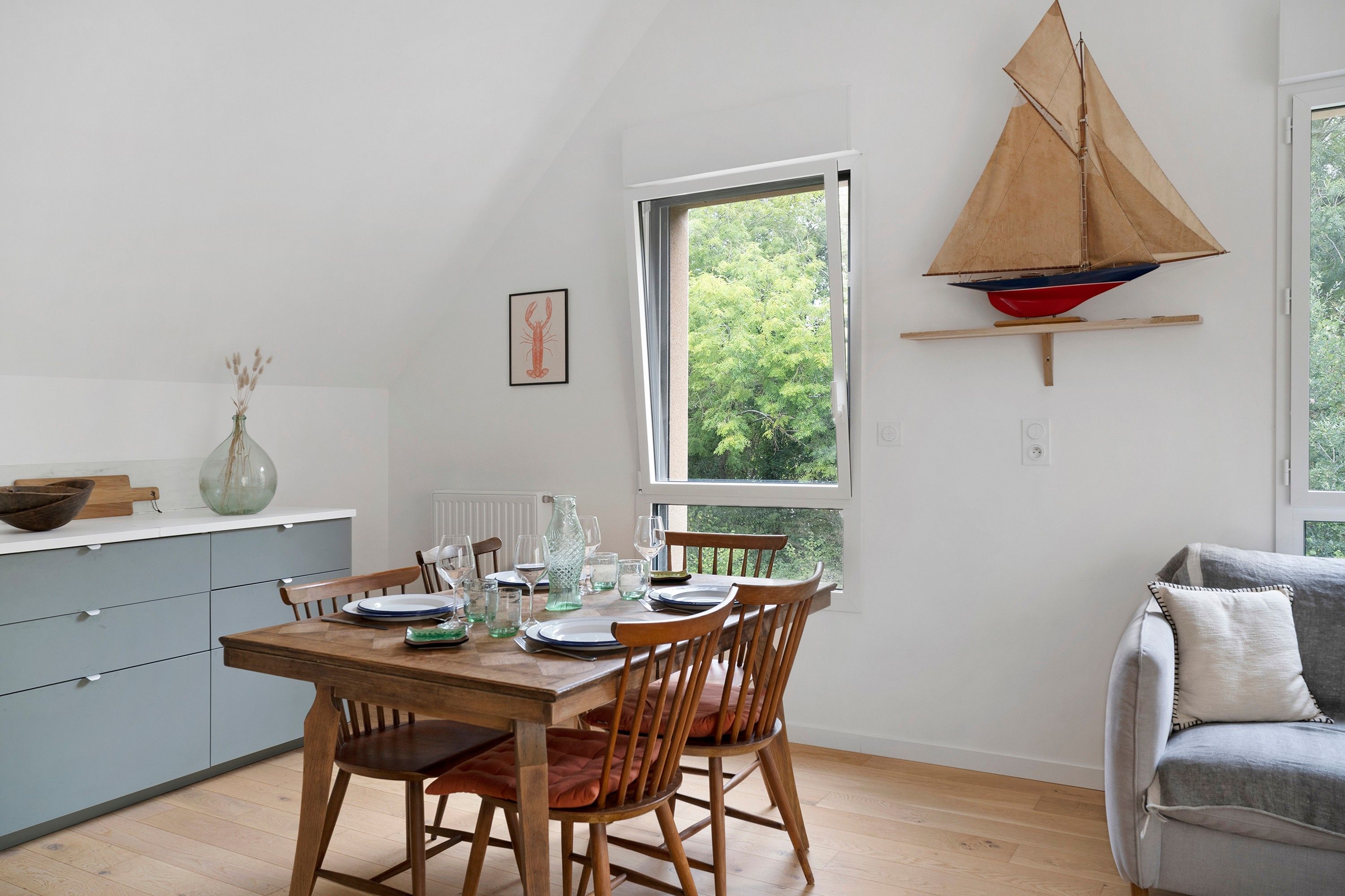 A dining area with a wooden table set for four, a grey sofa, and a sailboat model on a shelf.