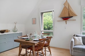 Een eetgedeelte met een houten tafel voor vier personen, een grijze bank en een model zeilboot op een plank.