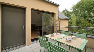 Une terrasse extérieure avec une table dressée pour le repas, des chaises vertes et une porte-fenêtre coulissante donnant sur un espace de salle à manger intérieur.