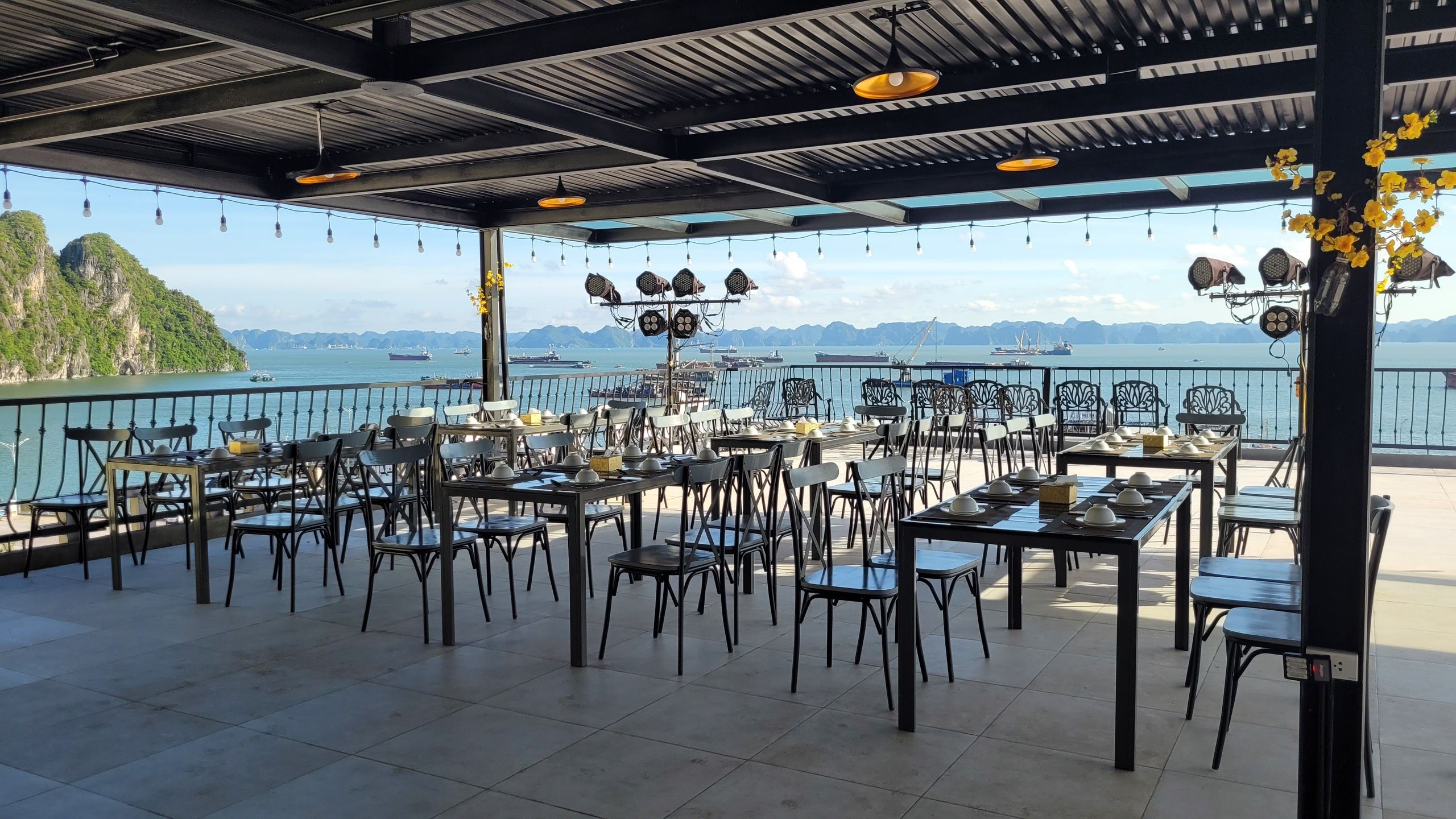 Un área de comedor al aire libre con mesas y sillas, con vista a un cuerpo de agua donde se ven barcos a lo lejos.