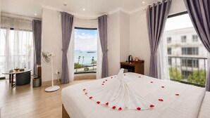 A hotel room with a large bed, a view of the sea, and a balcony with a table and chairs. - The Confetti Hotel Ha Long (Hong Gai)