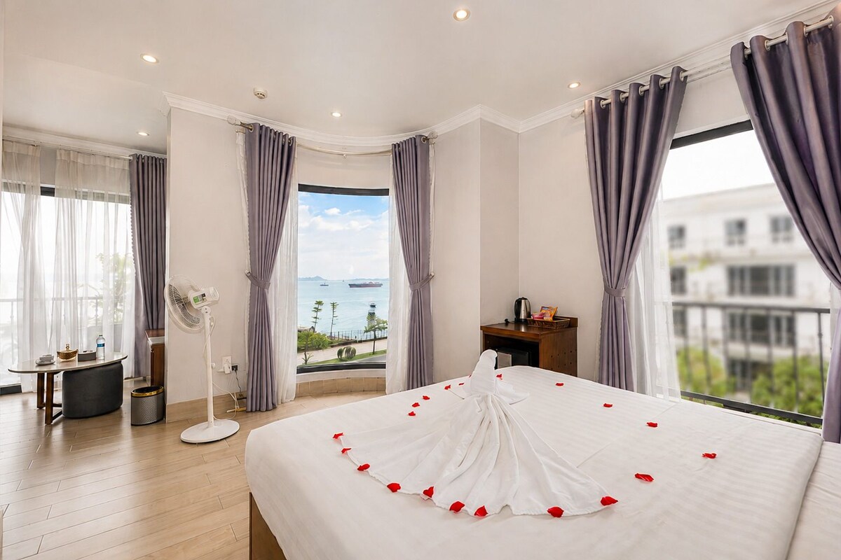 Habitación de hotel con una cama grande, vista al mar y balcón con mesa y sillas.